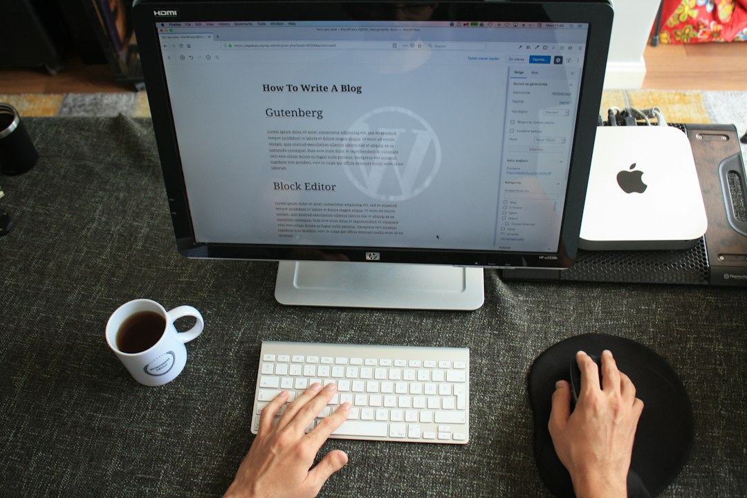ある人が机の上で白いキーボードを打ちながら、大きなモニターでWordPressホスティングに関するブログ記事の書き方を編集しています。コーヒーのカップ、マウスパッド、テクノロジーアクセサリーがきちんと近くに並べられている。